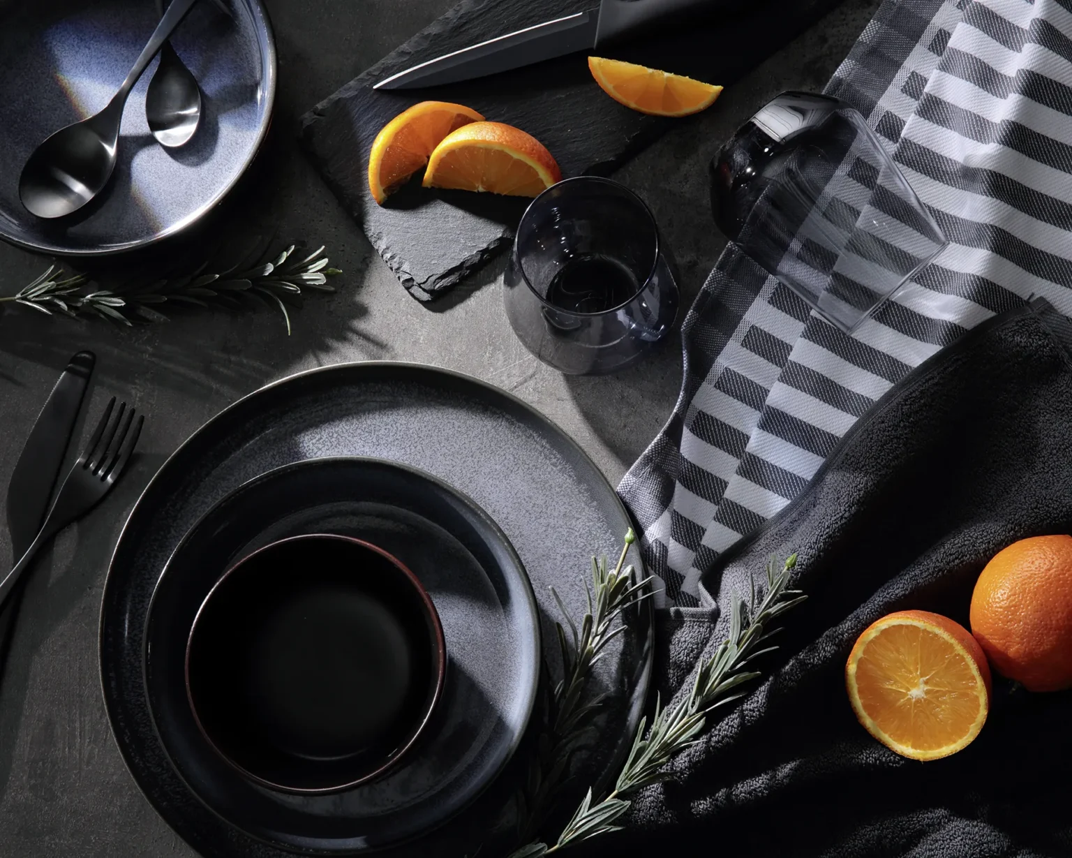 Ein gedeckter Tisch mit Orangen und Rosmarinzweigen. Ein Glas ist umgefallen.